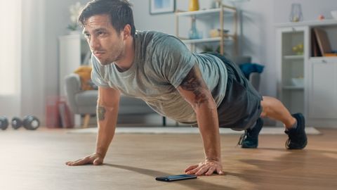 Athletic Fit Man in T-shirt and Shorts is Doing Push Up Exercises While Using a Stopwatch on His Phone. He is Training at Home in His Living Room with Minimalistic Interior.