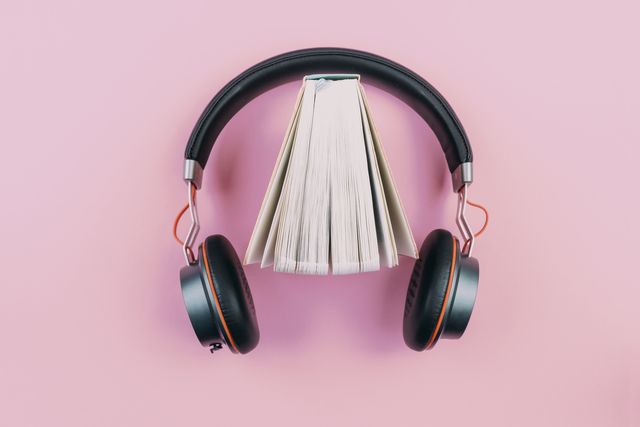 audiobook concept, top view of stereo headphones and fanned out hardcover book on pink desktop