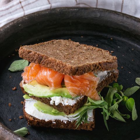Avocado, salted salmon and ricotta rye sandwich with fresh herds on black clay tray with checkered kitchen towel over dark wooden textured background. Healthy eating theme.