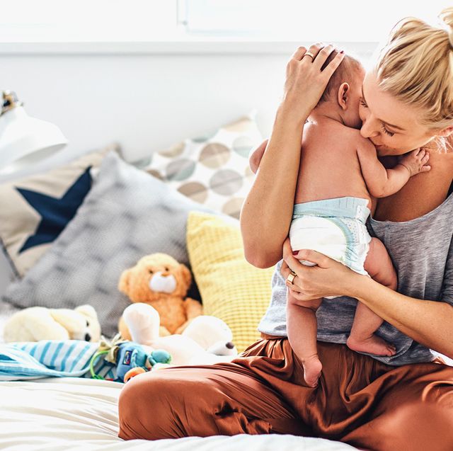 mother sitting on bed with pillows and stuffed animals holding and kissing baby in diaper