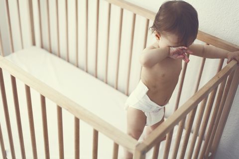 una bebé niña, de pie en su cuna, se restriega los ojos vestida con un pañal