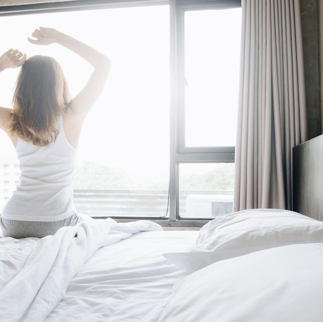 Back view of woman raised hands up and stretching in bed after waking up in the morning, start a new day with happiness.