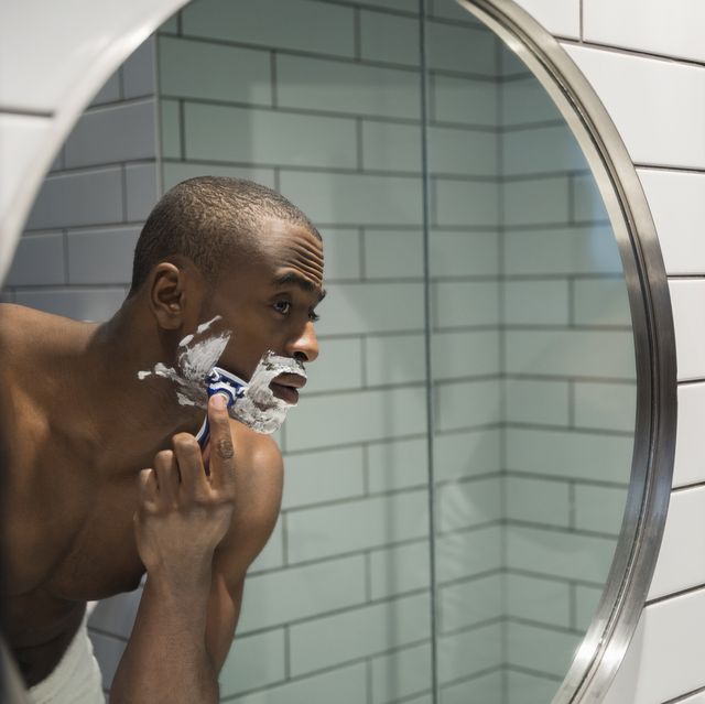 Bare chested man shaving face in bathroom mirror