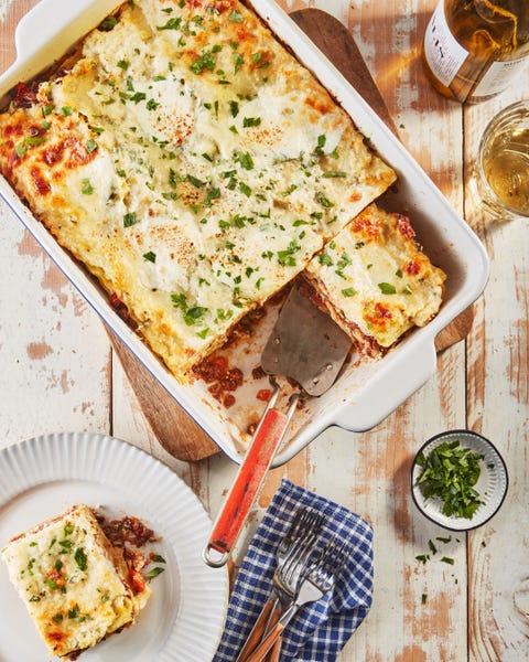 lasagna bolognese in a white rectangle casserole dish with a red serving spatula and a portion of lasagna on a white plate next to the dish