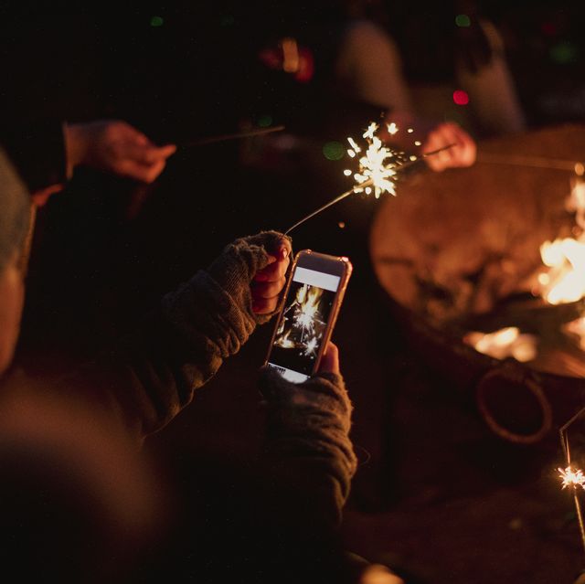 woman is using her smart phone to take a photo of a sparkler she is holding by the bonfire