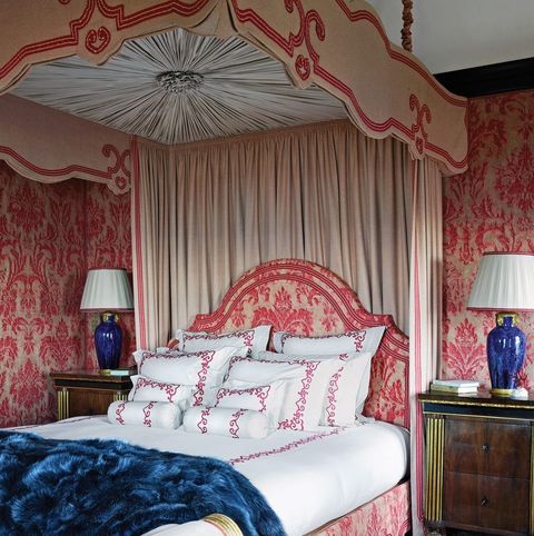 lush bed with curtains and scalloped headboard baseboard upholsterd in red floral that matches wallpaper behind bed is made with white sheets and stacks of pillows with red embroidered trim a