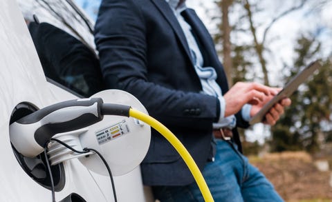 business man standing near charging electric car or ev car and using tablet in the street