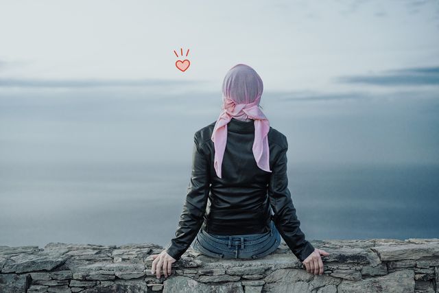mujer con pañuelo rosa en la cabeza sentada en un muro frente al mar, cáncer de mama