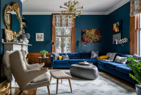 large living room with dark blue walls and tufted l shaped dark blue sofa spanning two walls with a silver triangular coffee table at center and large artwork pieces hanging on the walls a white fireplace and chair face the sofa