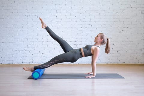 Caucasian young woman in grey sports suit doing pilates workout on math with fitness roller, loft style background, toned light blue