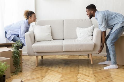 cheerful afro spouses placing couch furnishing empty room in new house