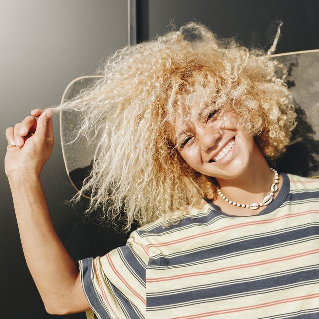 cheerful woman holding skateboard while pulling curly blond hair against wall