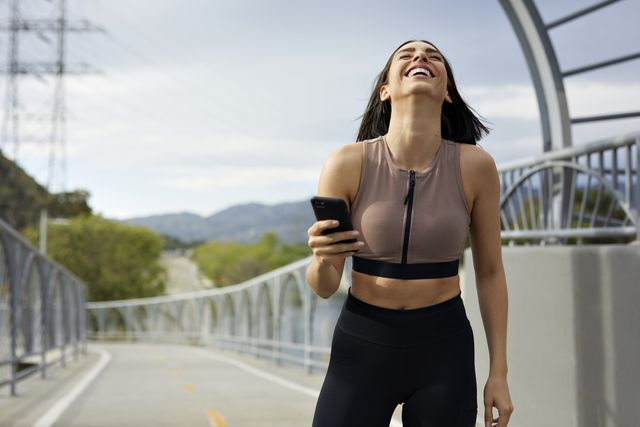 cheerful woman in sportswear with mobile phone standing on bridge