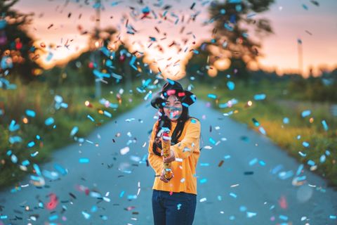 cheerful woman with colorful confetti standing on road during sunset