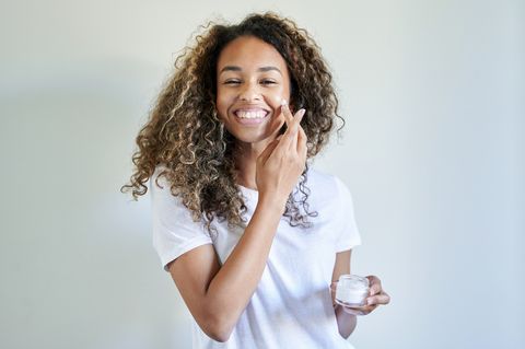 cheerful young woman applying facial cream against white wall