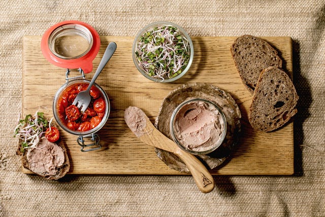 een plank met sneetjes brood, belegd met paté en een potje gedroogde tomaatjes