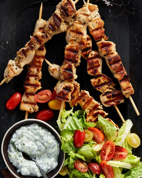 chicken souvlaki skewers surrounded by a bowl of tzatziki, cherry tomatoes, and chopped romaine on a black platter