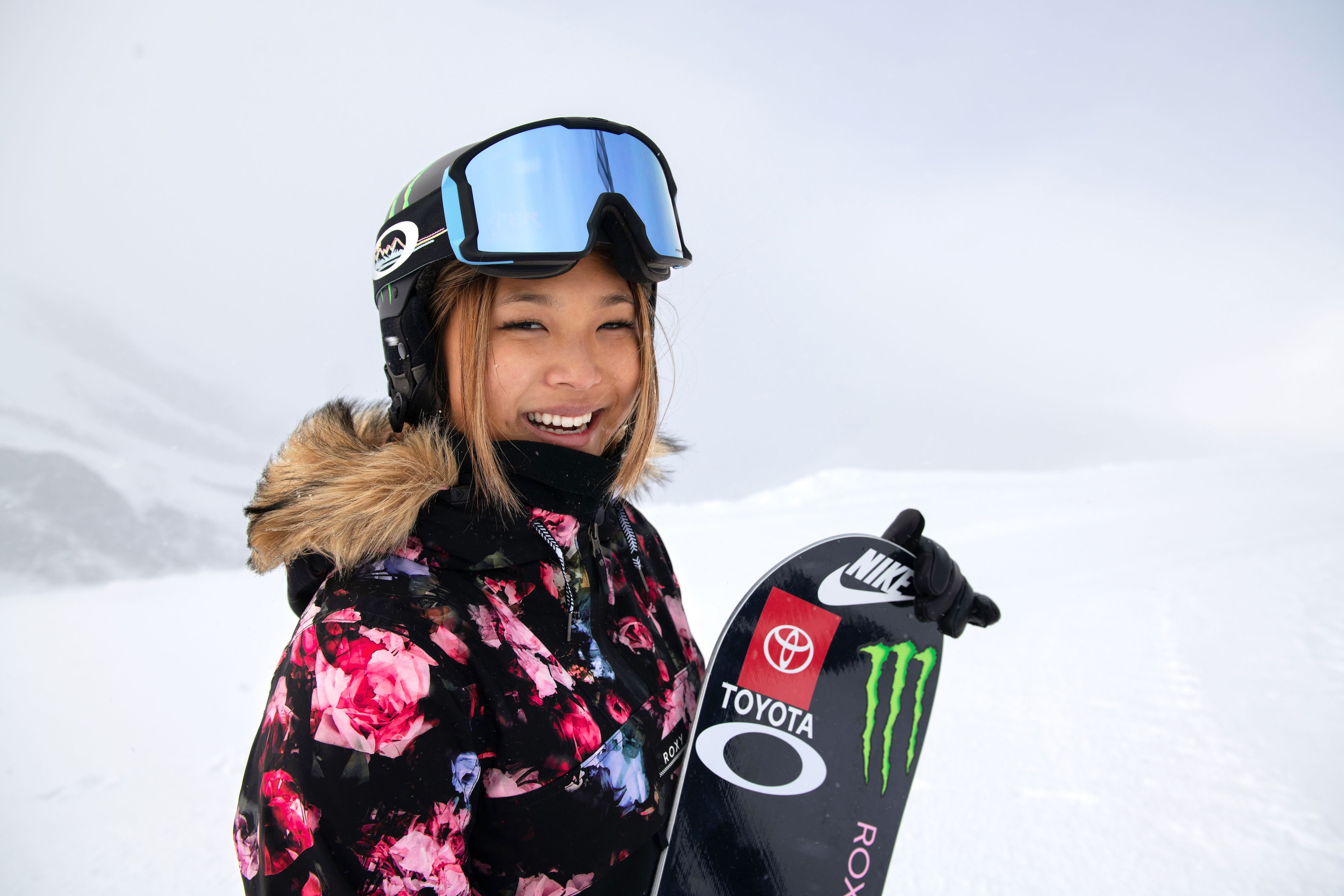 Snowboarder Chloe Kim standing on a snow covered hill holding her snowboard up smiling for the camera.