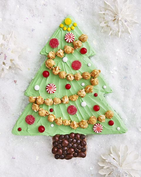 a sheet cake cut into the shape of a christmas tree with brown frosting for the stump and green frosting for the leaves and caramel corn for garland and carious white and red candies for ornaments