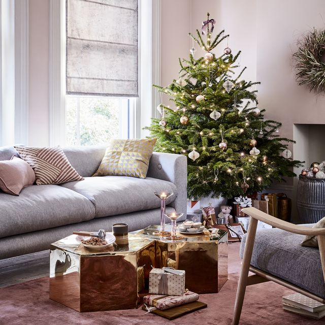 christmas tree origin, why do we have christmas trees, christmas tree in the corner of a lliving room grey sofa and gold hexagon coffee tables, light pink walls and grey roman blindsreflected glory stunningmirroredbrass tables, bronzeandpinkbaublesandaccentsofgoldbringamodernopulence toarelaxedlivingroom