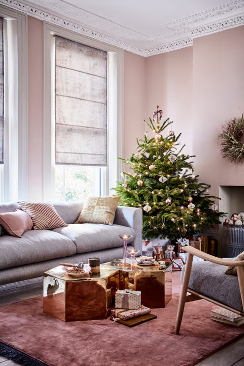 christmas tree in the corner of a lliving room grey sofa and gold hexagon coffee tables, light pink walls and grey roman blindsreflected glory stunningmirroredbrass tables, bronzeandpinkbaublesandaccentsofgoldbringamodernopulence toarelaxedlivingroom