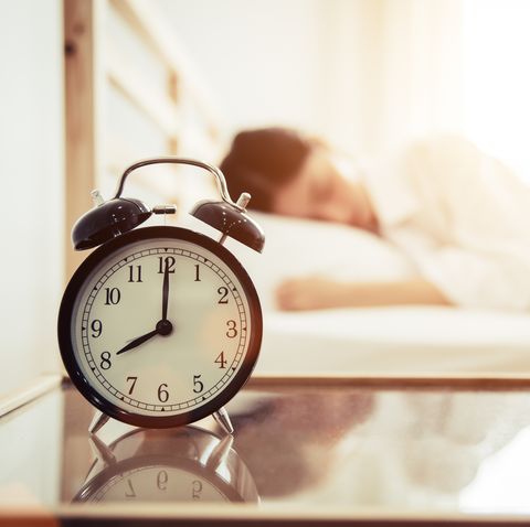 Close-Up Of Alarm Clock On Table By Woman Sleeping On Bed At Home