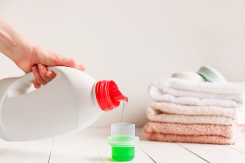 close up of female hands pouring liquid laundry detergent into cap on white rustic table with towels on background in bathroom