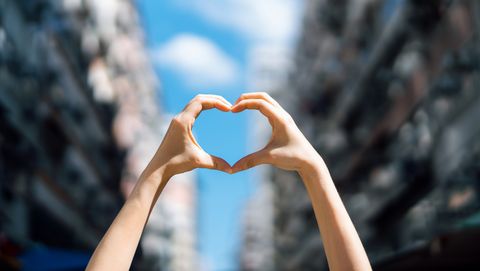 close up of woman's hand raised and making a heart shape over clear blue sky in the city