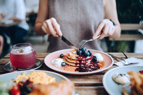 close up of young woman sitting at dining table eating pancakes with blueberries and whipped cream in cafe, with english breakfast and french fries served on the dining table eating out lifestyle