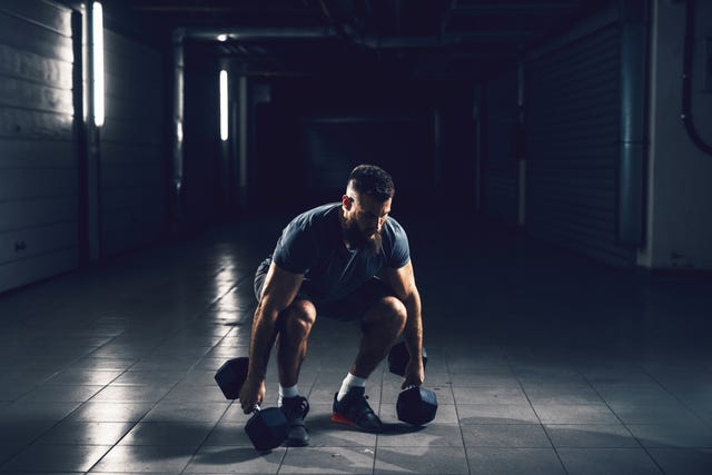 Close up view of focussed hardworking active fitness strong muscular bearded bodybuilder man crouching before raising heavyweight dumbbell in the underground garage or urban gym while looking far away. close up view of focussed hardworking active fitness strong muscular bearded bodybuilder man crouching before raising heavyweight dumbbell in the underground garage or urban gym while looking far away