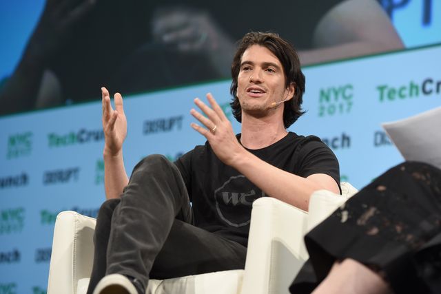 wework founder adam neumann, a white man, sits in a black t shirt and pants in a white chair on a stage