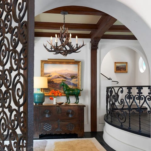 A List 2022 - ELLE DECOR entry type of area with platform to the right that leads upstairs through an archway and beautiful wrought iron heart shaped railings around the platform and on the gate and a soft colored rug on the wood floors