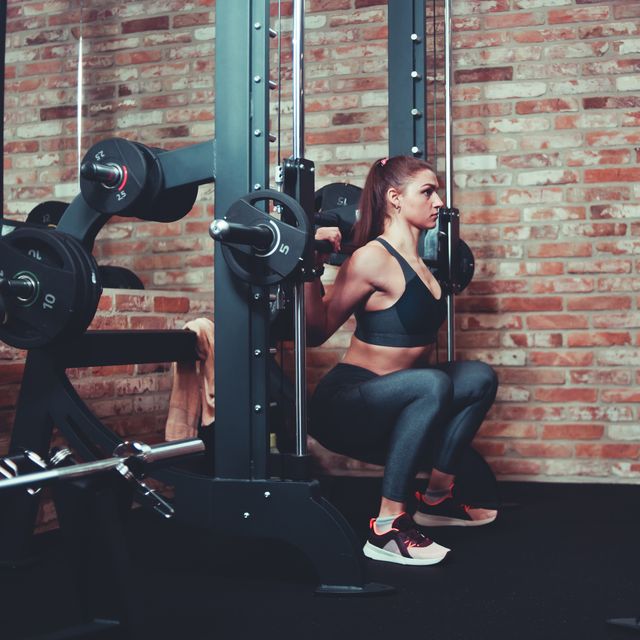 concentrated athletic woman exercising squatting at smith machine rack bar against brick wall in gym