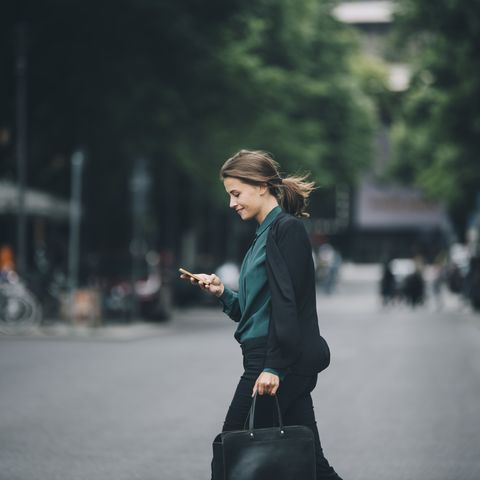 confident businesswoman using smart phone while crossing street in city
