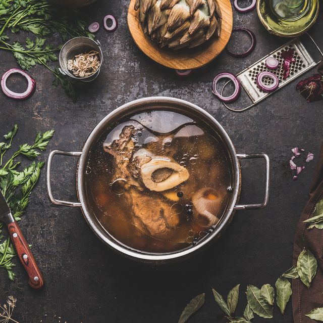 cooking pot with beef bone broth or stock on kitchen table with ingredients