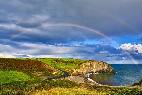 copper coast, ireland with road winding through green fields and a bay below a cliiff over it all is rainbow
