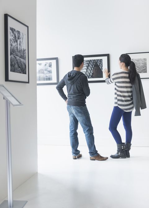 man and woman admiring framed photographs in gallery, she is gesturing toward a photo and has her coat casually slung over her shoulder