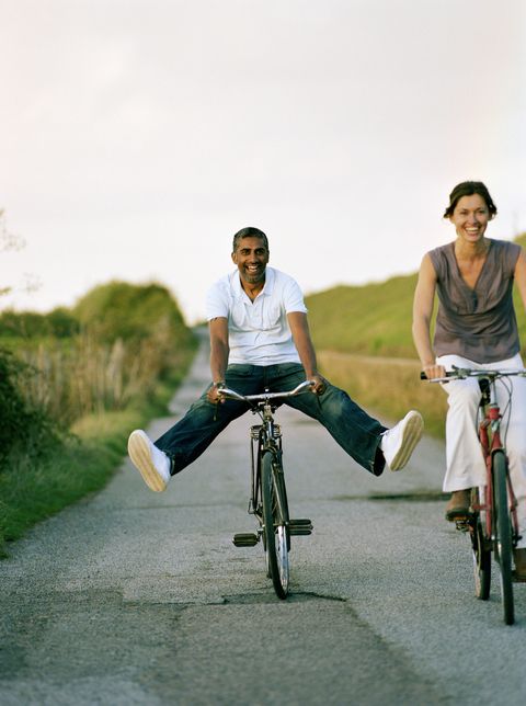 couple riding bicycles on paved path cutting through a field, man balancing with outstretched legs