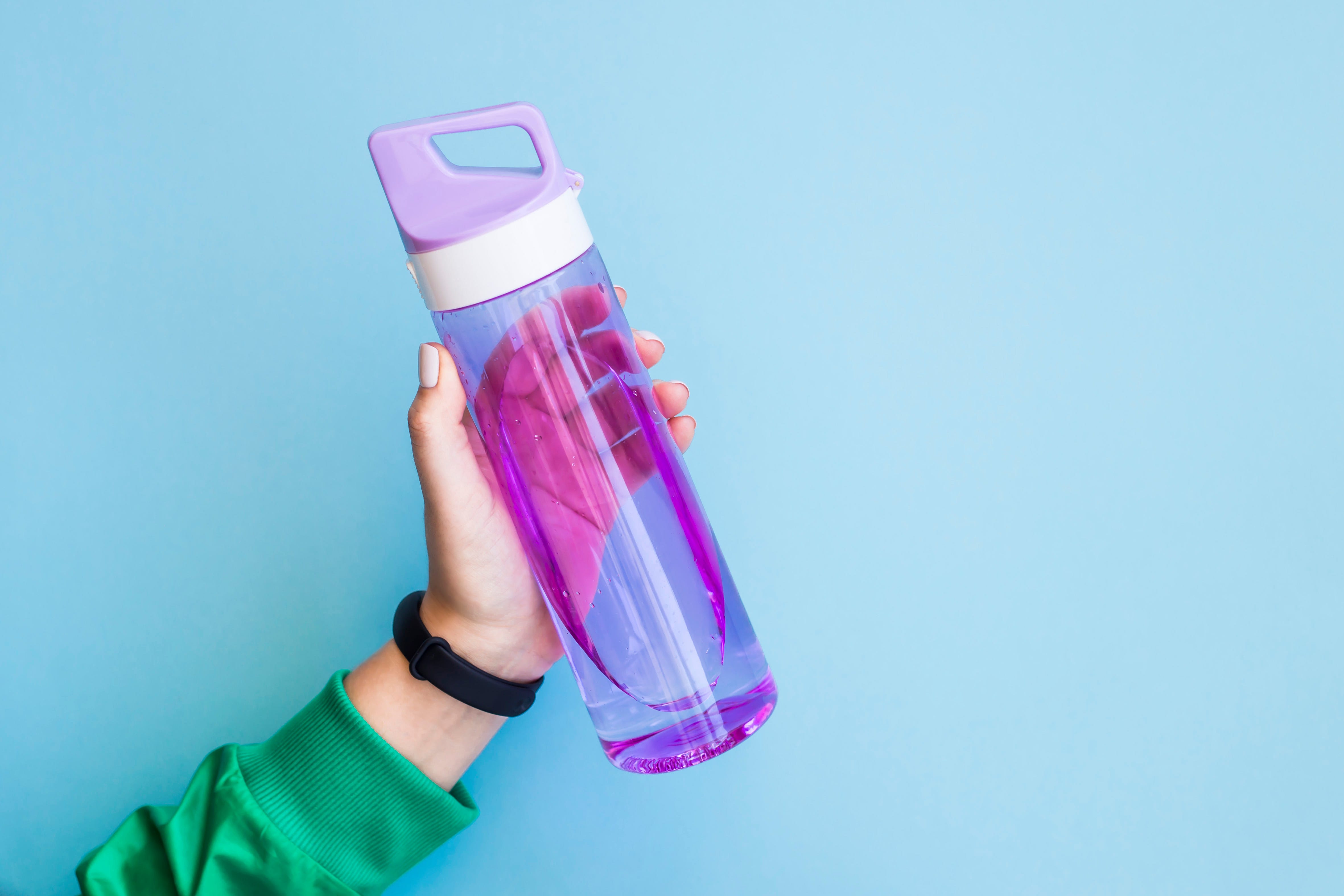 mano recortada de mujer sosteniendo una botella de agua púrpura contra un fondo azul