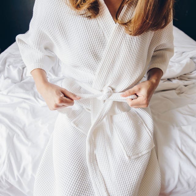 cropped shot view of young woman sitting on the bed and tying up belt of white bathrobe