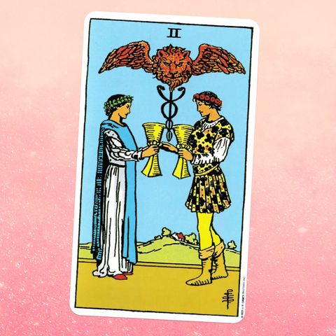 the Two Cups tarot card, showing a woman in a white robe and a man in a yellow page outfit facing each other, holding goblets, with a winged lion above them