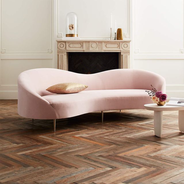 pale pink, sofa with a curve in the middle of its back, sitting on a wood floor in front of a fireplace there is a small white table in front of it with flowers on it
