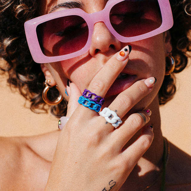 close up portrait of a fashionable tattooed woman with curly hair, colorful rings, sunglasses and trendy nail art, posing in front of an orange background