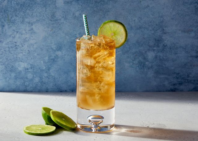 glass of a dark and stormy cocktail with a paper straw and a lime sitting on a stone surface with a marbled blue background