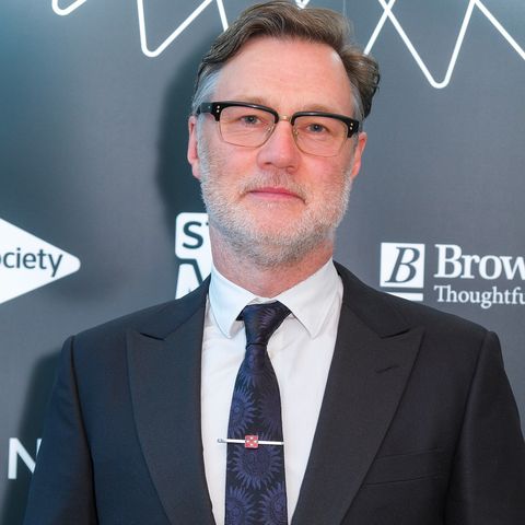 david morrissey, an older man looks at the camera with a neutral facial expression, he wears glasses with graying hair slicked back, black suit jacket, white shirt and blue tie