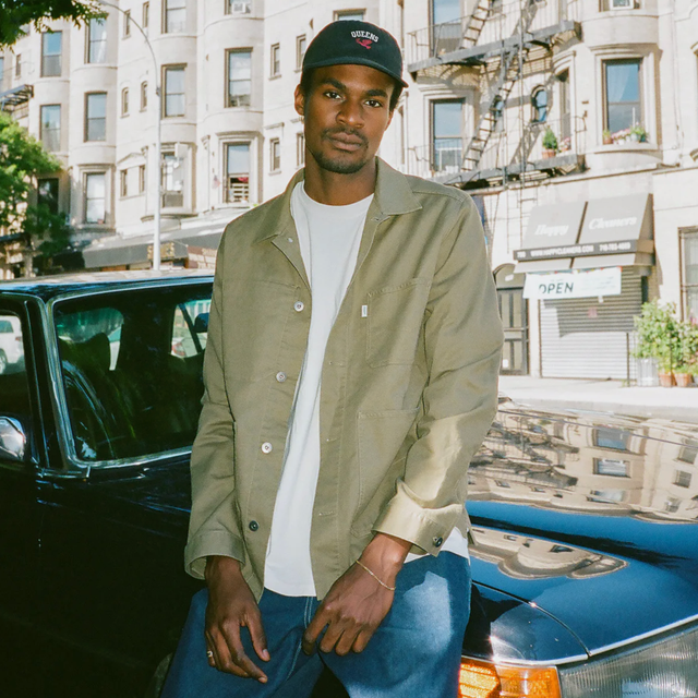 Wellen Stretch Chore Coat a model sitting on a car wearing a black hat, white t shirt, olive chore coat and blue jeans
