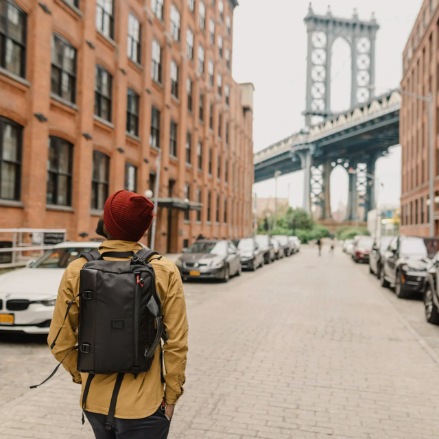 Topo Designs Commuter Briefcase a model walking down a street in brooklyn towards the manhattan bridge