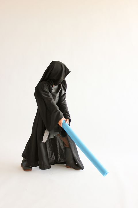 boy in an anakin skywalker costume with black hooded robe, black vest, holding blue pool noodle as light saber