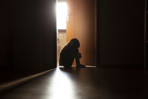 despairing child sitting with head on knees in the dark frame of a doorway, backlit by a room behind flooded with daylight