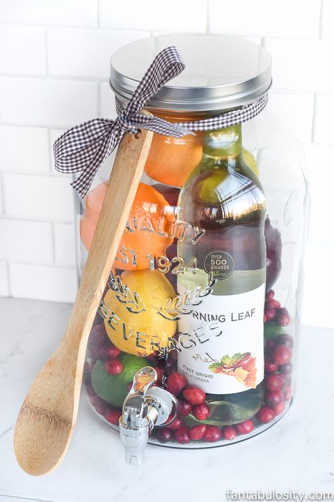large glass drink dispenser filled with oranges, lemons, cranberries, and bottle of pinot grigio for making sangria, with wooden spoon tied around lid with gingham ribbon